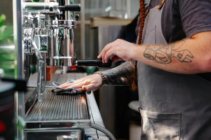 Barista with tattoos preparing coffee on an espresso machine, using a towel to clean the portafilter