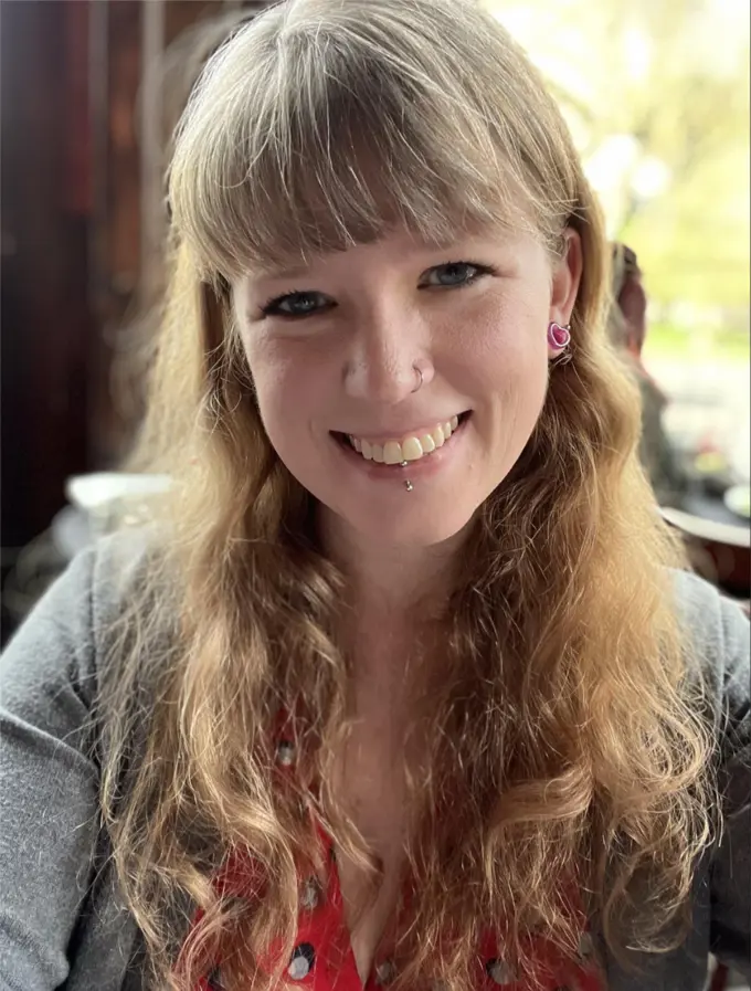 Profile Picture of Janae Cram. A smiling woman with long blonde hair, a nose ring, and a lip piercing, wearing a red patterned top and gray sweater. She is seated in a warmly lit environment with a natural outdoor background