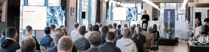 A large room with a full audience, listening to the speaker on stage. There are 2 screens showing the presentation. The room is bright and welcoming.