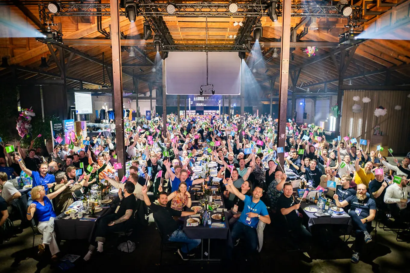 A large group of people seated at tables in a spacious, well-lit venue, holding up colorful cards and smiling for the camera.