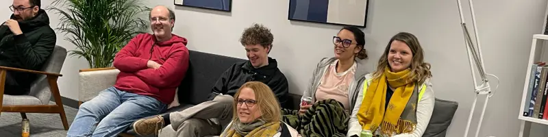 6 people are sitting together around a couch, lots of smiles.