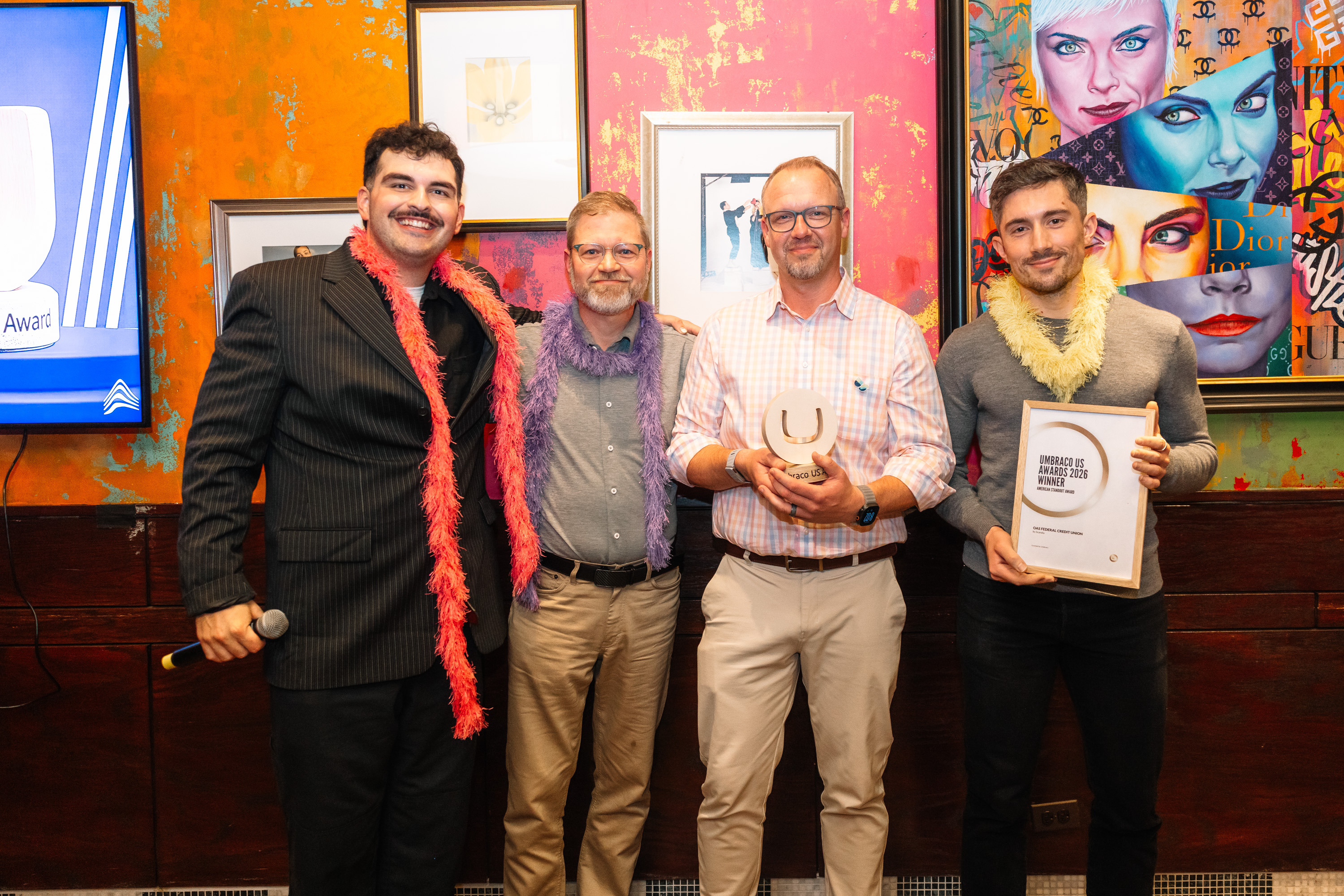 Four men posing indoors at an awards event, holding trophies and a framed certificate; colorful wall art behind.