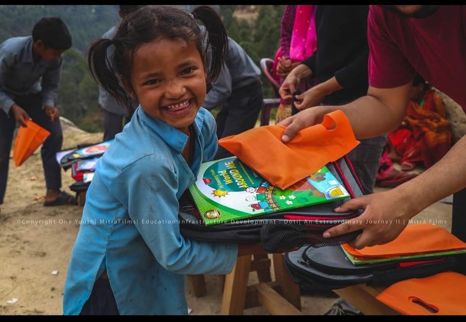 A young girl with pigtails smiles brightly as she is handed a school backpack stacked with a colorful "World Around Me" textbook and an orange pouch.