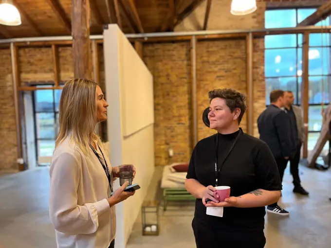 Two women standing and talking in a modern, rustic styled room, holding drinks, with one in a white blouse and black trousers and the other in a black turtleneck, both wearing conference badges.