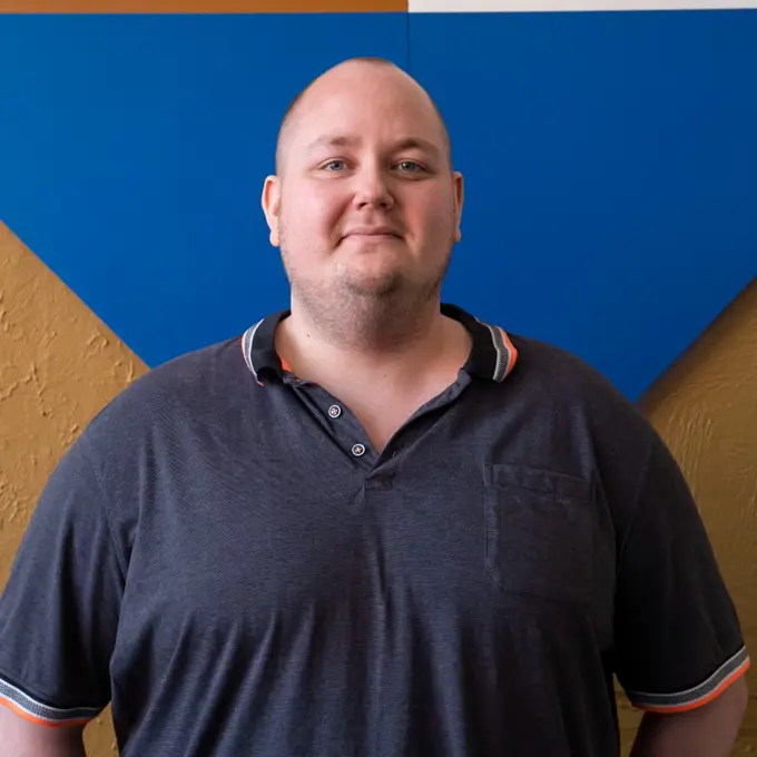 Headshot of Mikkel, a white man with stubble wearing a dark blue shirt against a blue and gold background.