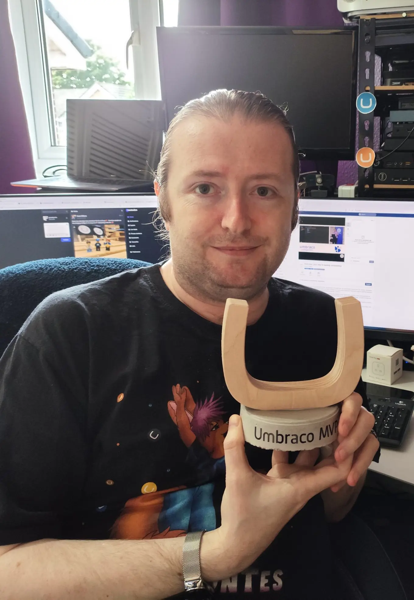 Image of Terence Burridge, wearing a black tshirt, holding his wooden U shaped Umbraco MVP award, in front of his home office setup