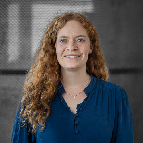 Smiling red-haired woman with blue blouse, posed in front of a gray studio backdrop.