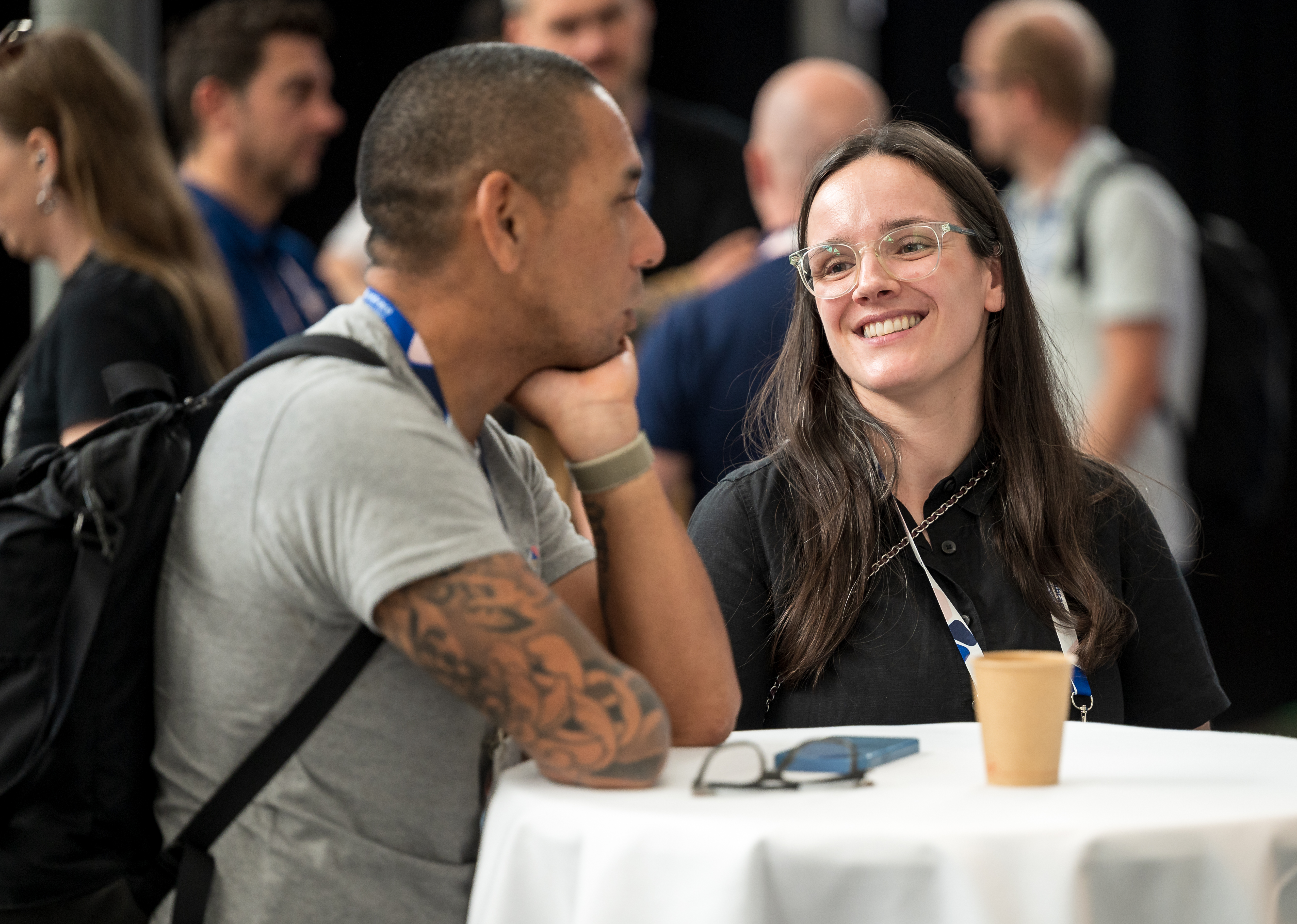 Woman with glasses smiles at a conference table while seated next to a man; other attendees are in the background.