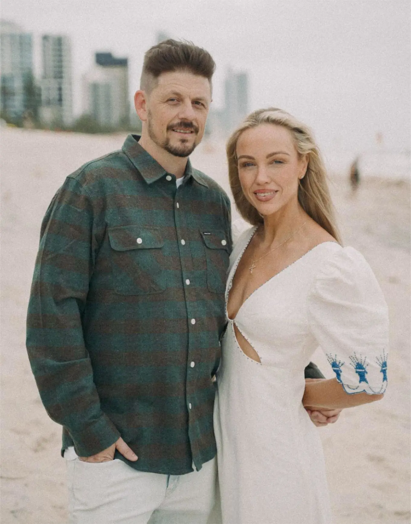 Image of a couple, Pete and Cherie Gregory, on a beach. Pete wearing pale chinos and a checked shirt. Cherie wearing a white, puff sleeve dress.