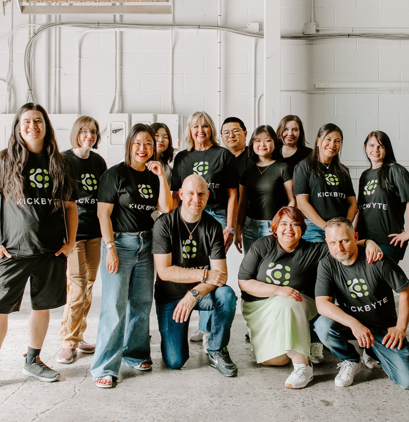 Group photo of coworkers wearing black “KICKBYTE” T-shirts with green paw-print logos in an industrial room