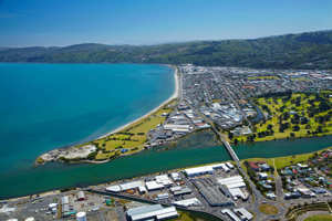 A high-angle aerial panoramic photograph showcasing an industrial and residential coastal town situated next to a bright blue bay and green hills.