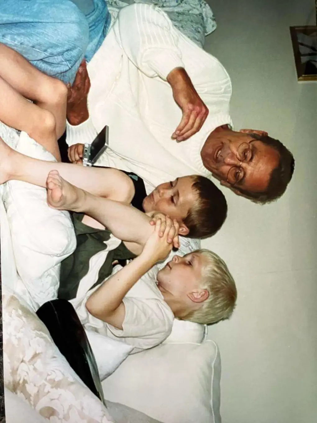 Three people, one older man with glasses and two young white boys, sitting on a couch playing on a handheld device.