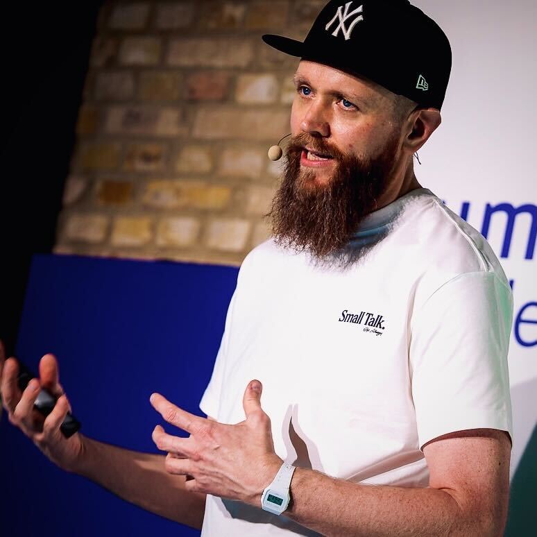 A man with a full beard and a black baseball cap speaks while gesturing with his hands during a presentation. He is wearing a white T-shirt with small text and a light-colored watch, standing in front of a brick wall and a blue banner.