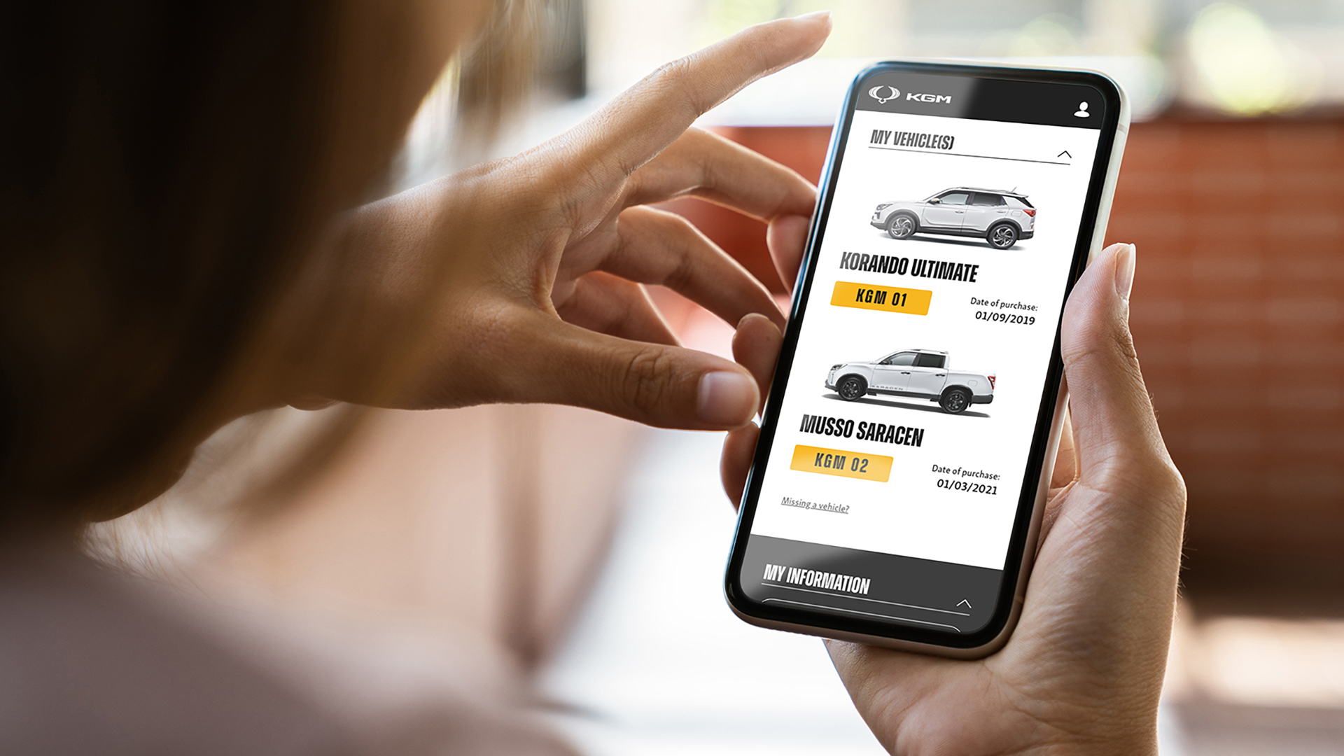 Hand holding a smartphone showing KGM “MY VEHICLES” with vehicle listings KGM 01 and KGM 02 and purchase dates