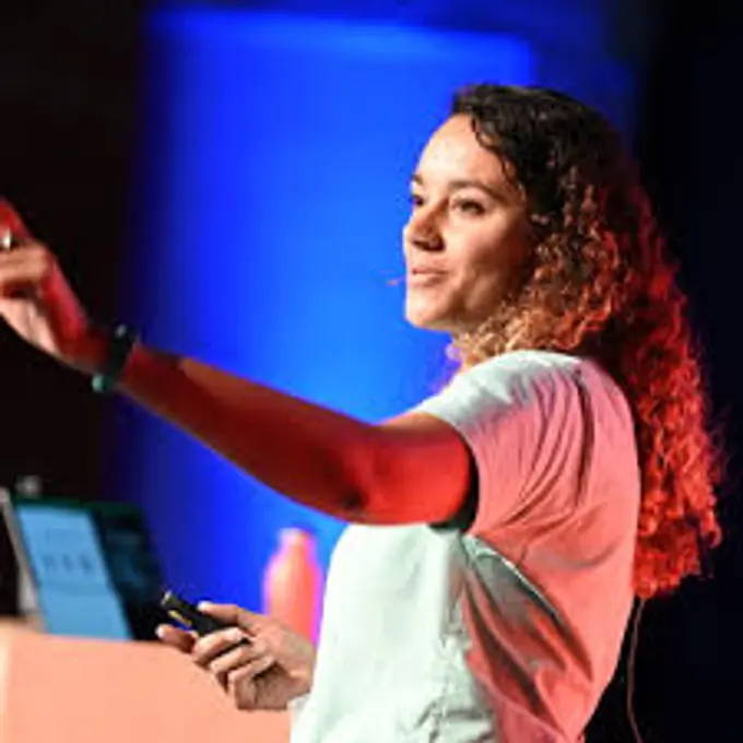 Profile Picture of Emma Burstow, in the spotlight at a conference, presenting on stage, set against a Blue light background, wearing a mint green tshirt, with long curly dark hair. 