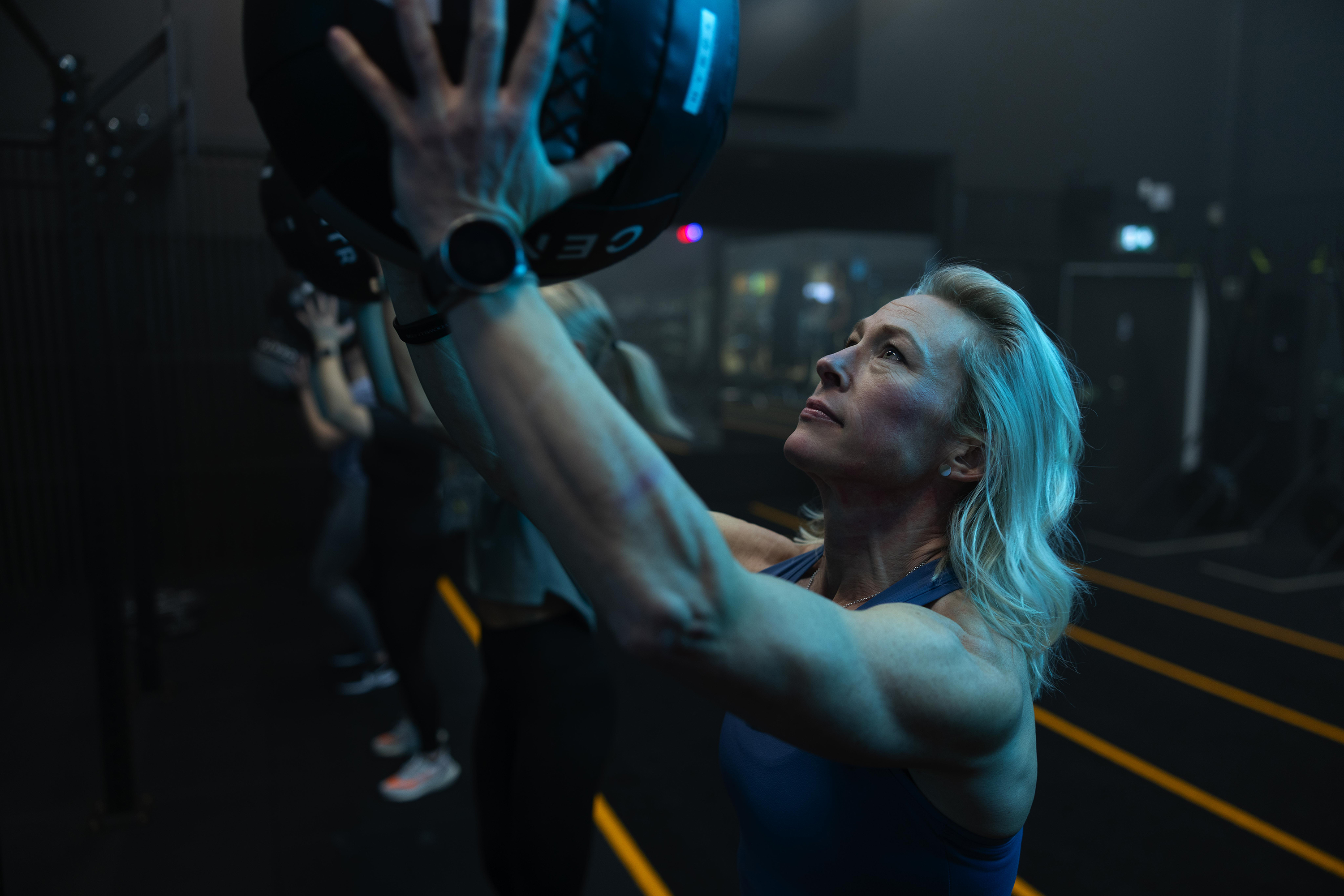 Woman with blue hair lifts heavy fitness equipment, focused upward in a dim gym.