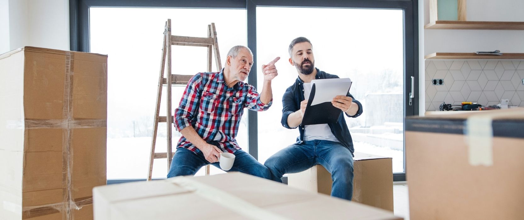 Two men in a room with moving boxes discussing plans; one points toward a window while the other holds a clipboard.