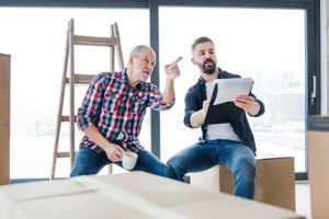 Two men in a room with moving boxes discussing plans; one points toward a window while the other holds a clipboard.
