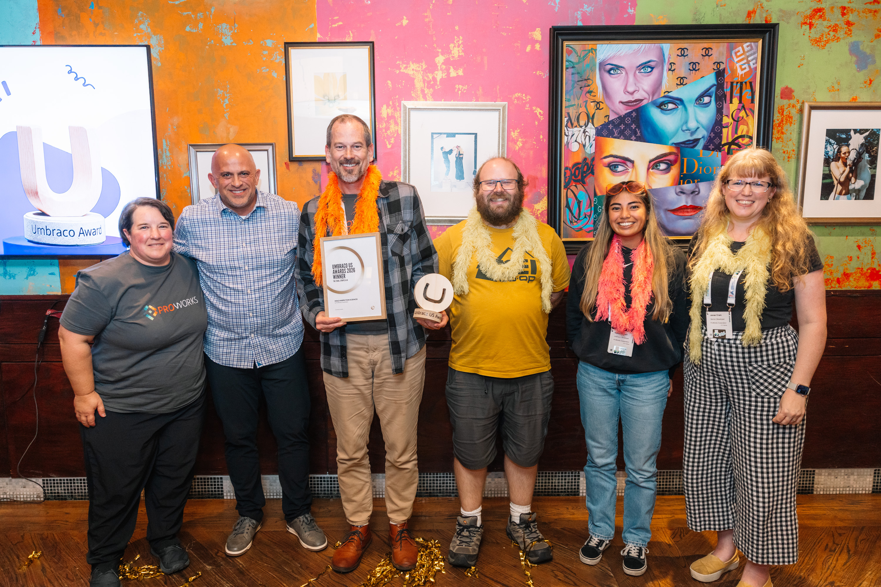 Group of six people at an award event, holding certificates and wearing colorful feather boas, with framed artwork behind them