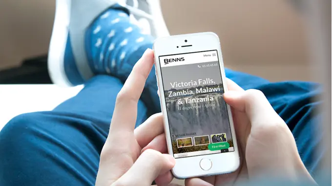 A person sitting with their feet up, holding a white phone in their hands. The phone displays BENN's website.