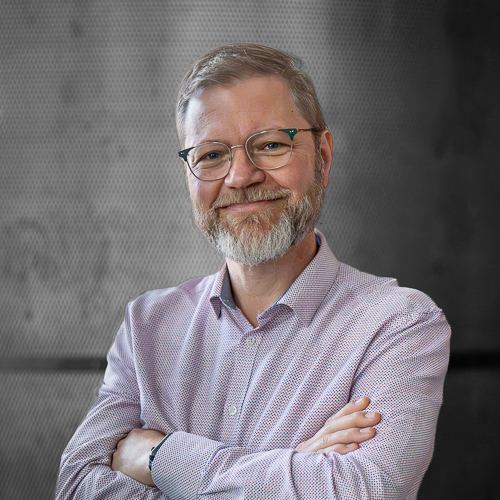 Smiling older man with gray hair, beard, and glasses wearing a pink checkered shirt with arms crossed.