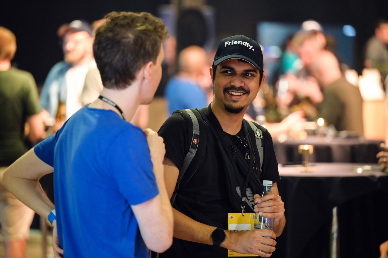 A smiling man in a black "Friendly." cap converses at a social gathering. He has a beard, wears a badge-laden black T-shirt, and carries a backpack. Holding a water bottle, he's likely at a conference, with other attendees and ambient lighting in the background.