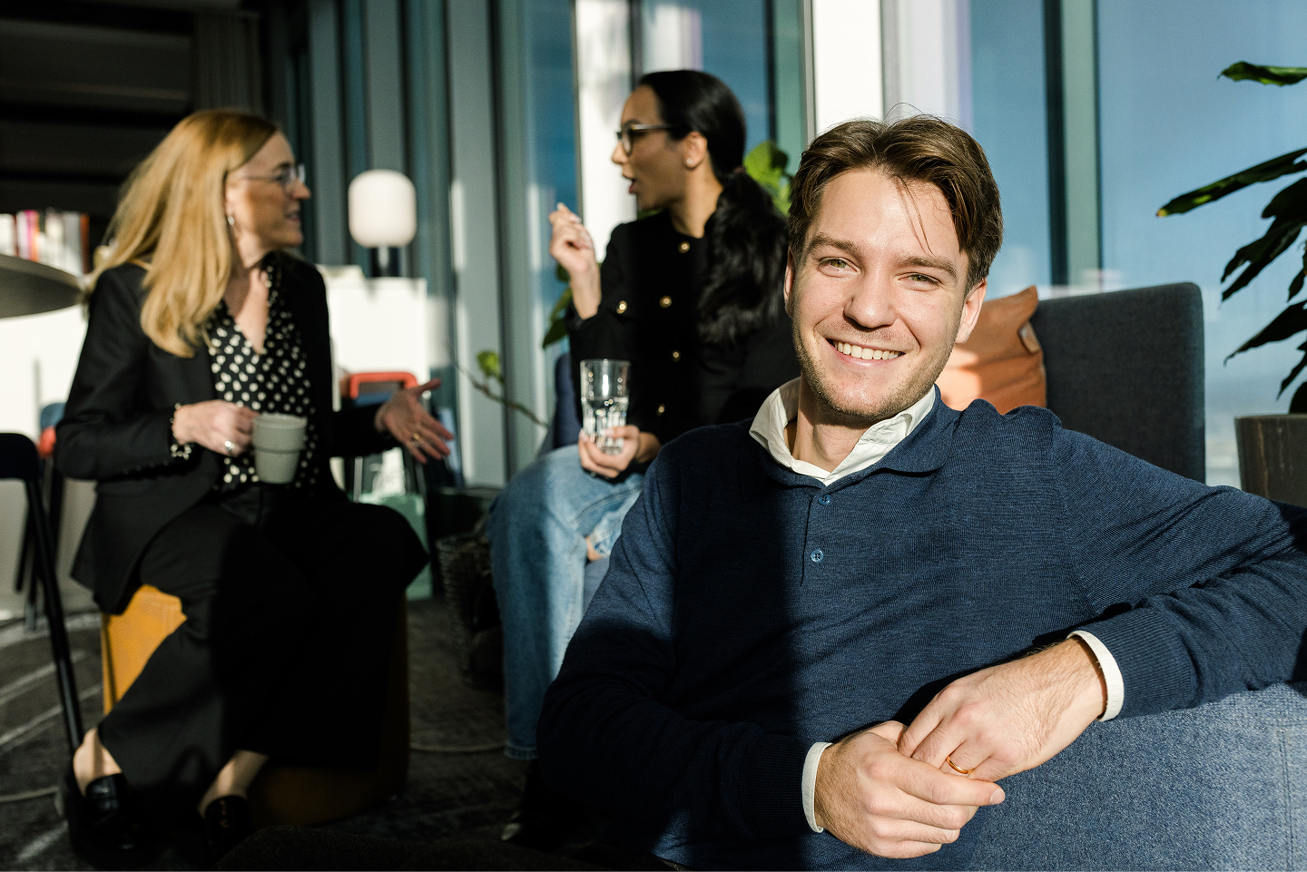 Smiling man seated in office with colleagues chatting in the background, holding a drink