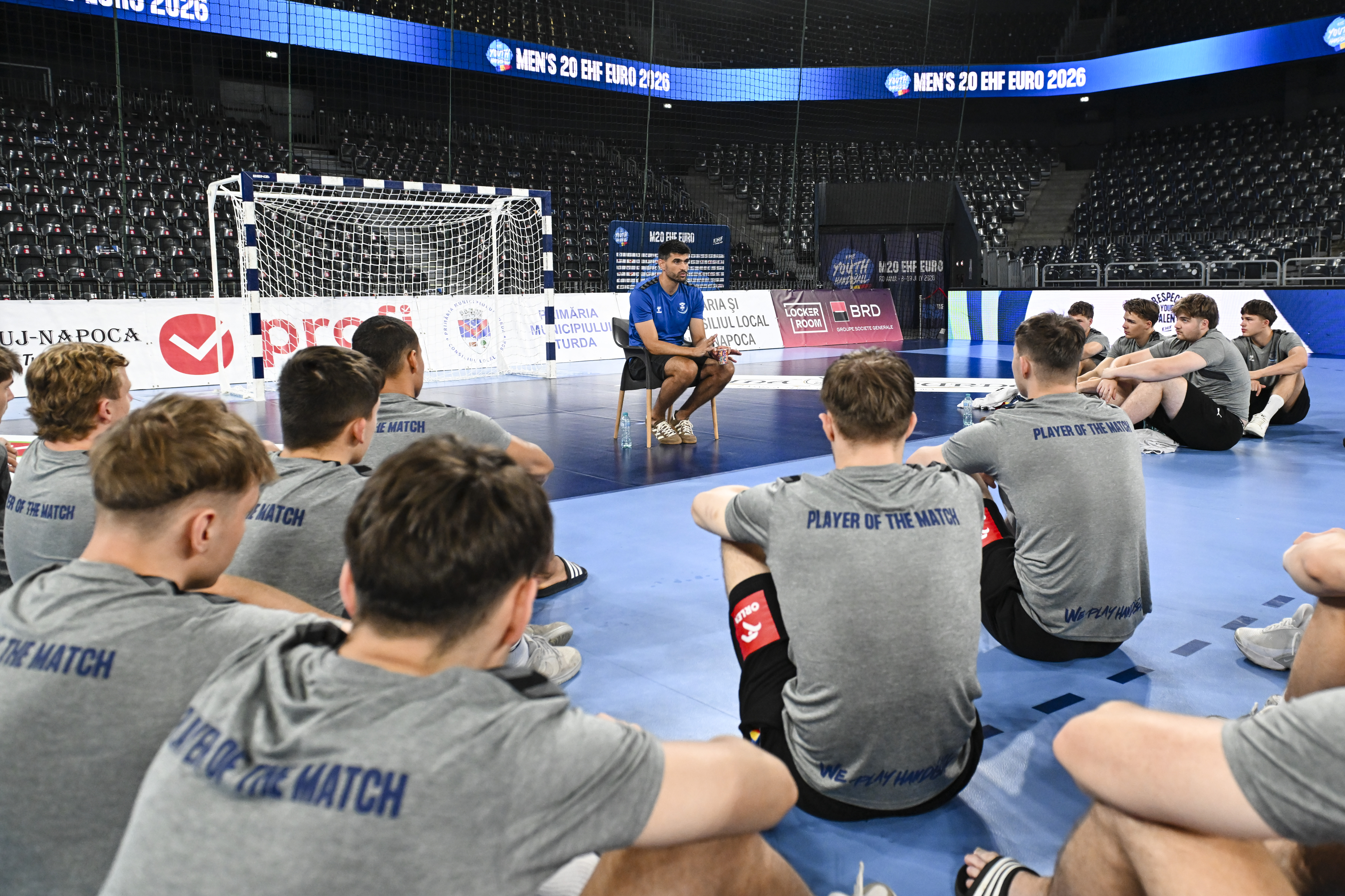 Men's 20 EHF EURO 2026 handball squad sitting on court during a coaching session with Player of the Match shirts.