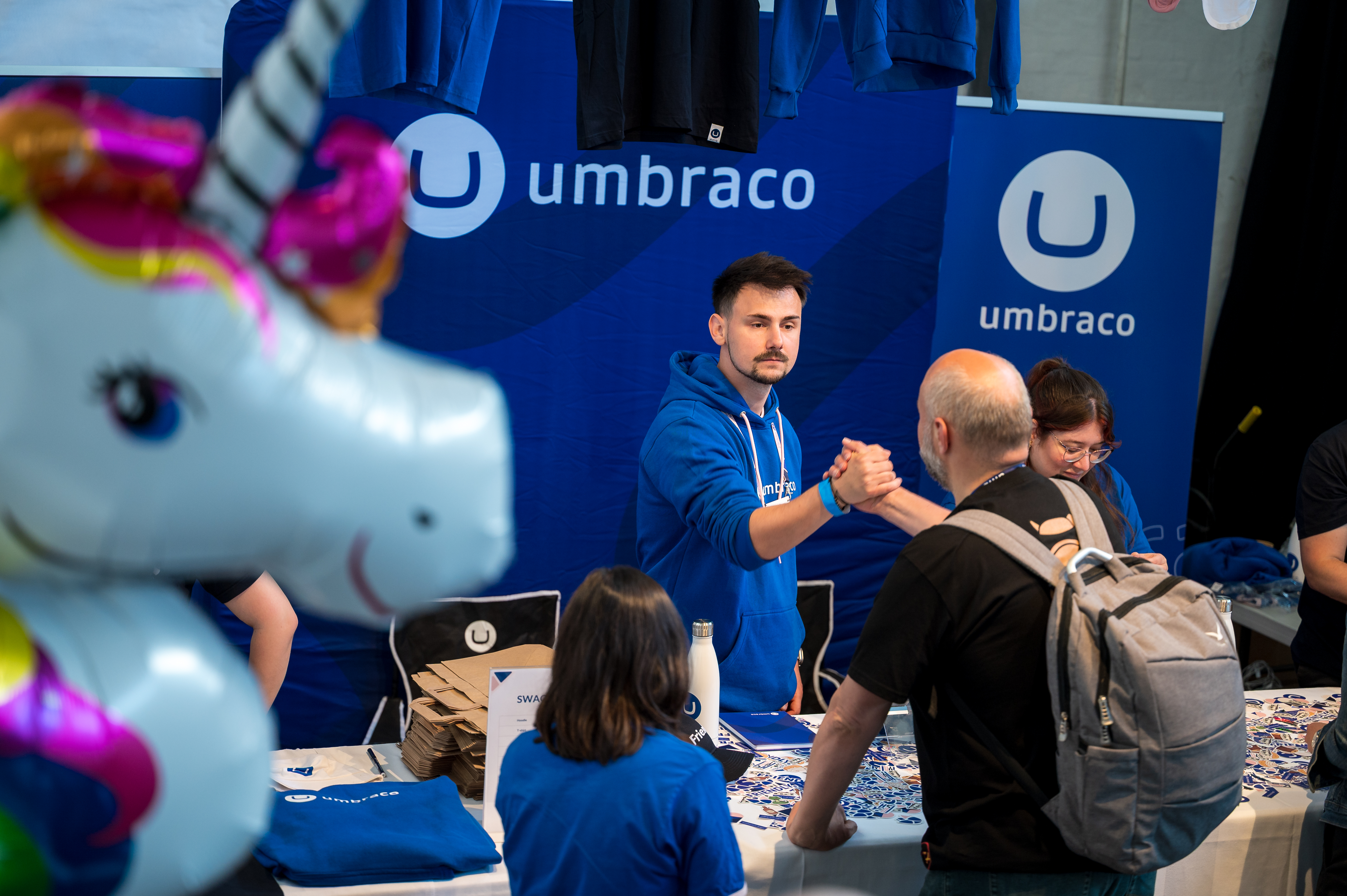 Man in blue hoodie shaking hands at trade booth with “umbraco” branding on blue banner