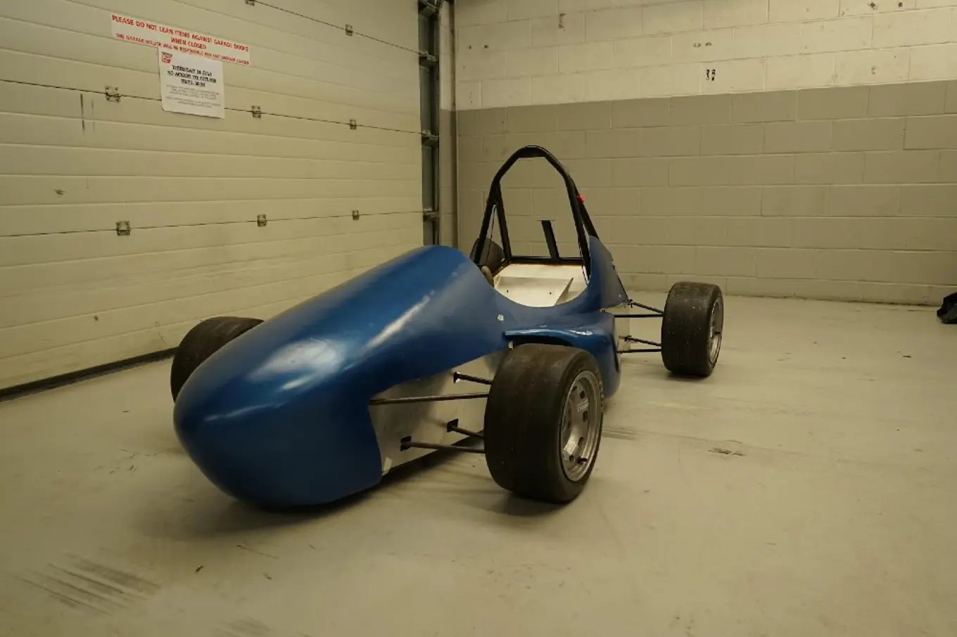 DIY racecar with a blue body and black wheels, shown in a beige brick room