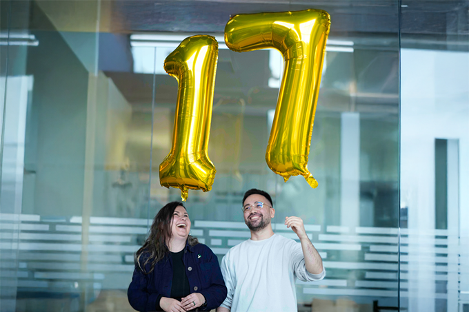 Two people smiling beneath large gold balloons shaped like the number 17