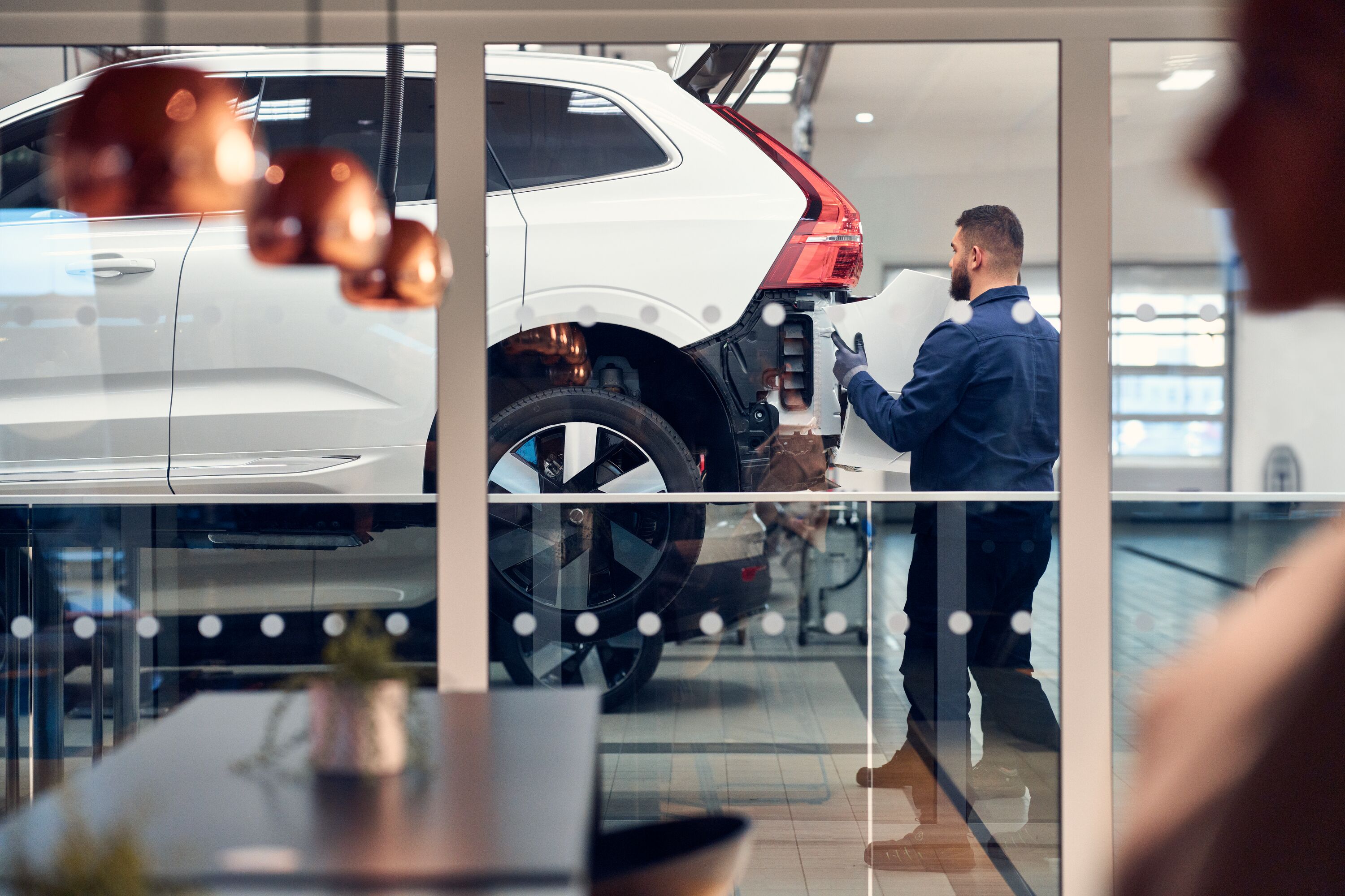 Viewed through a glass partition, a mechanic carries a white body panel toward the rear of a white Volvo SUV that is raised on a vehicle lift in a repair facility.