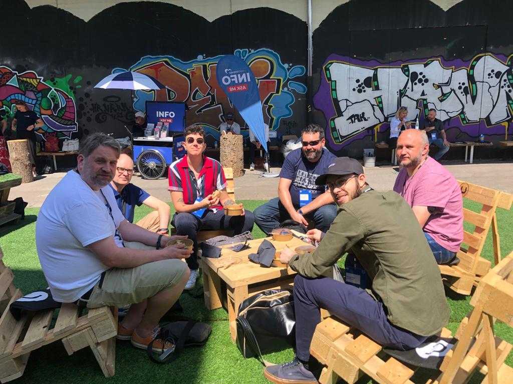 Group of people seated at wooden tables outside in front of graffiti mural, talking at an event booth.