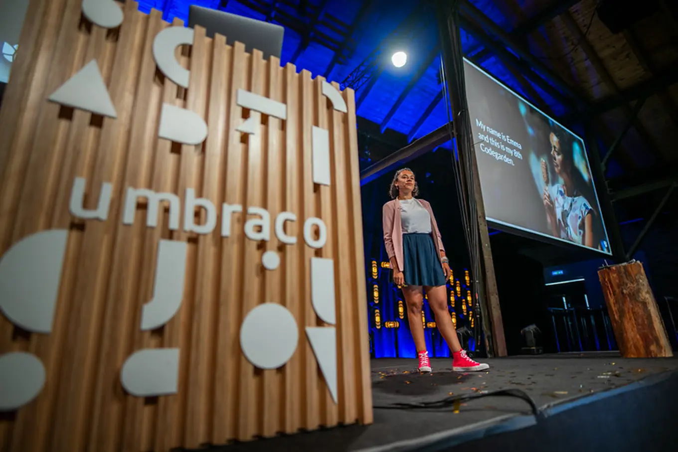 A woman standing confidently on a stage next to a podium with the "Umbraco" logo, delivering a presentation with a large screen behind her.