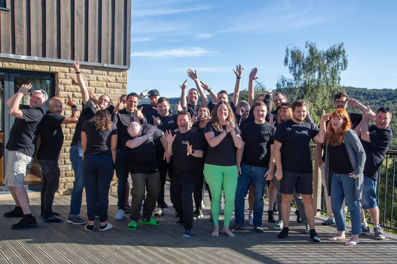 A group of people standing outdoors on a sunny day in front of a wooden building, all wearing black shirts. They are posing playfully, with arms raised, thumbs up, and some individuals striking fun, energetic poses. The group appears to be enjoying a team or community event, with a scenic background of trees and blue skies.