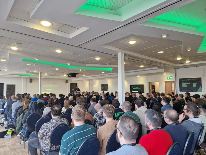 A presenter speaking at a tech conference in a well-lit room filled with seated attendees, with green highlights and branding of "UMBRACOSPARK" visible in the background.