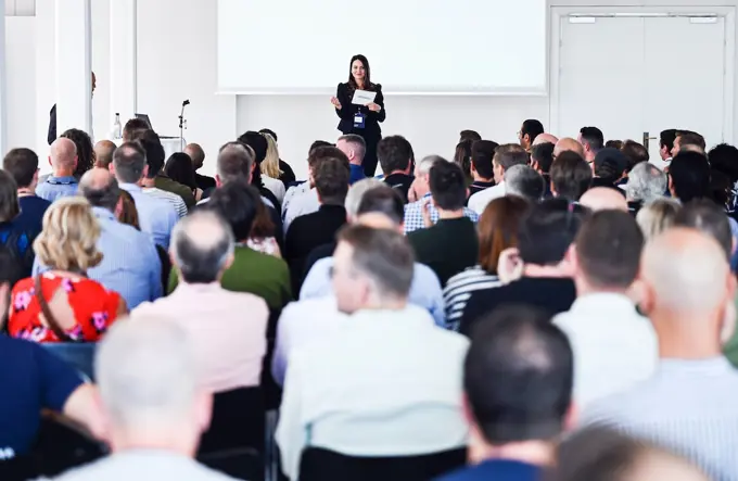 This image shows a large audience attentively watching a speaker presenting on stage in a bright, modern event space. The speaker is holding a microphone and appears to be addressing the group, with the audience's backs facing the camera. The setting suggests a professional conference or seminar. The diverse crowd is seated, indicating a formal presentation or talk in progress.