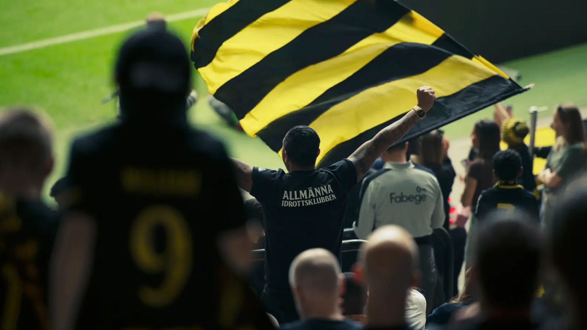 An AIK Fotboll supporter waving a black and yellow flag in the stands