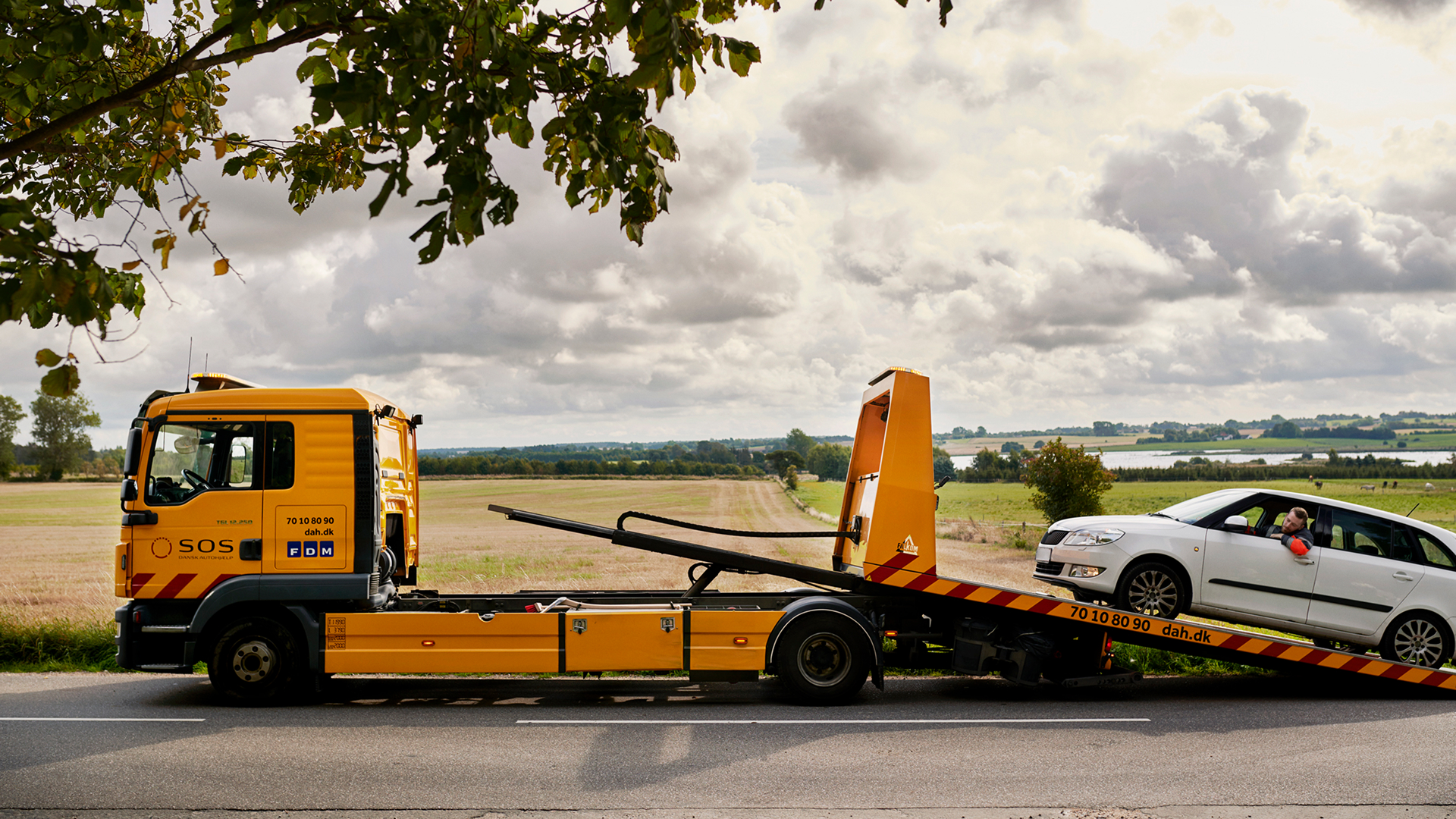 SOS Dansk Autohjælp roadside assistance tow truck loading a vehicle on a rural road, showing phone number 70 10 80 90 and website dah.dk.