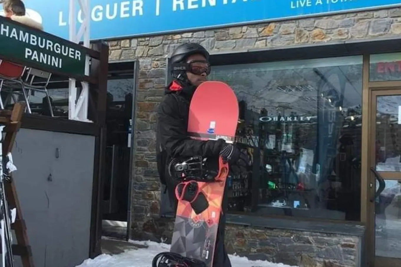 Myles is wearing a snowboarding helmet and glasses, and holding an orange-red snowboard