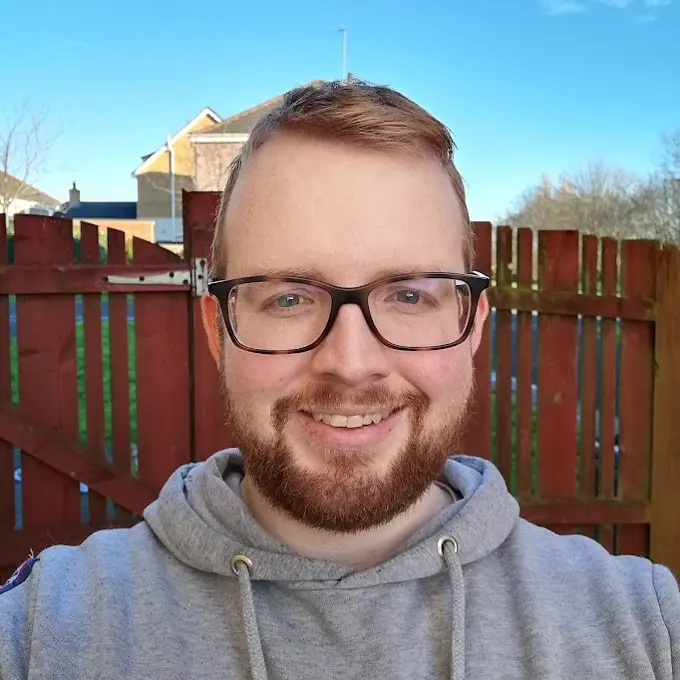 Mike is standing outside in front of a red painted fence. He has short ginger hair, glasses, a beard, and a big smile!