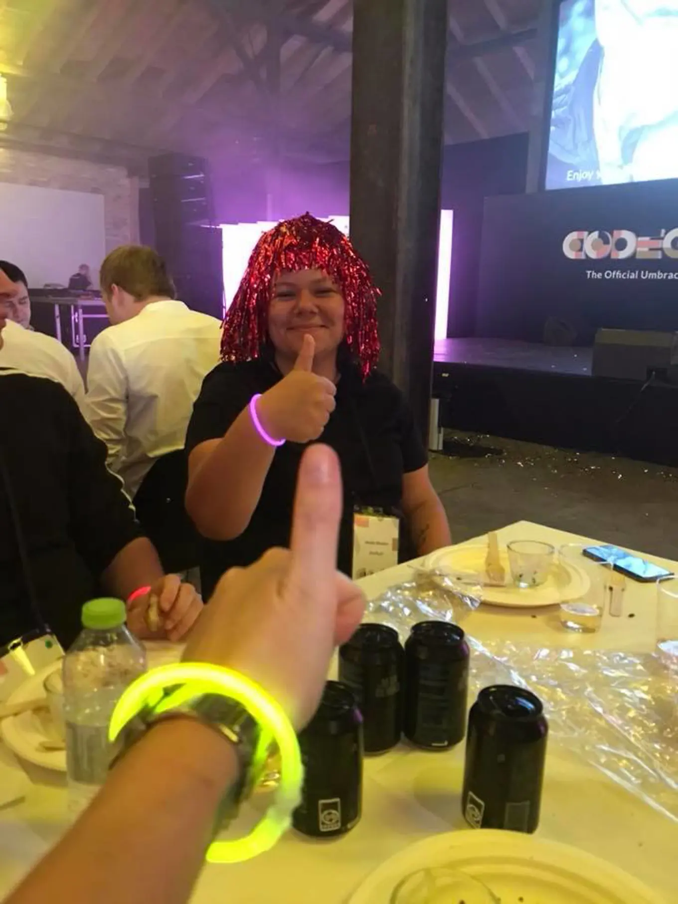 A smiling Amalie Wowern sat at a table at the Codegarden Dinner, giving a thumbs up while wearing a neon pink glowstick bracelet and a red, shiny tinsel wig.