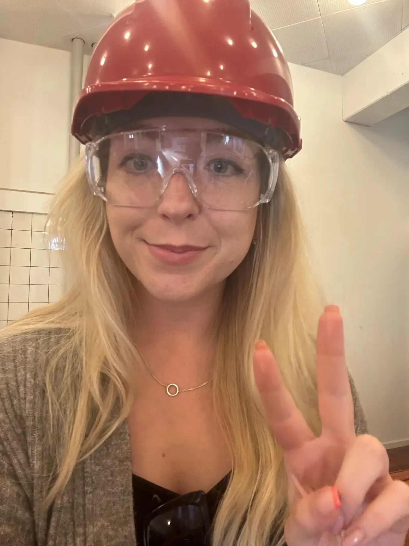 Image of Amy Czuba visiting Fjernvarme Fyn, wearing a red hard hat and safety goggles, while making a peace sign.