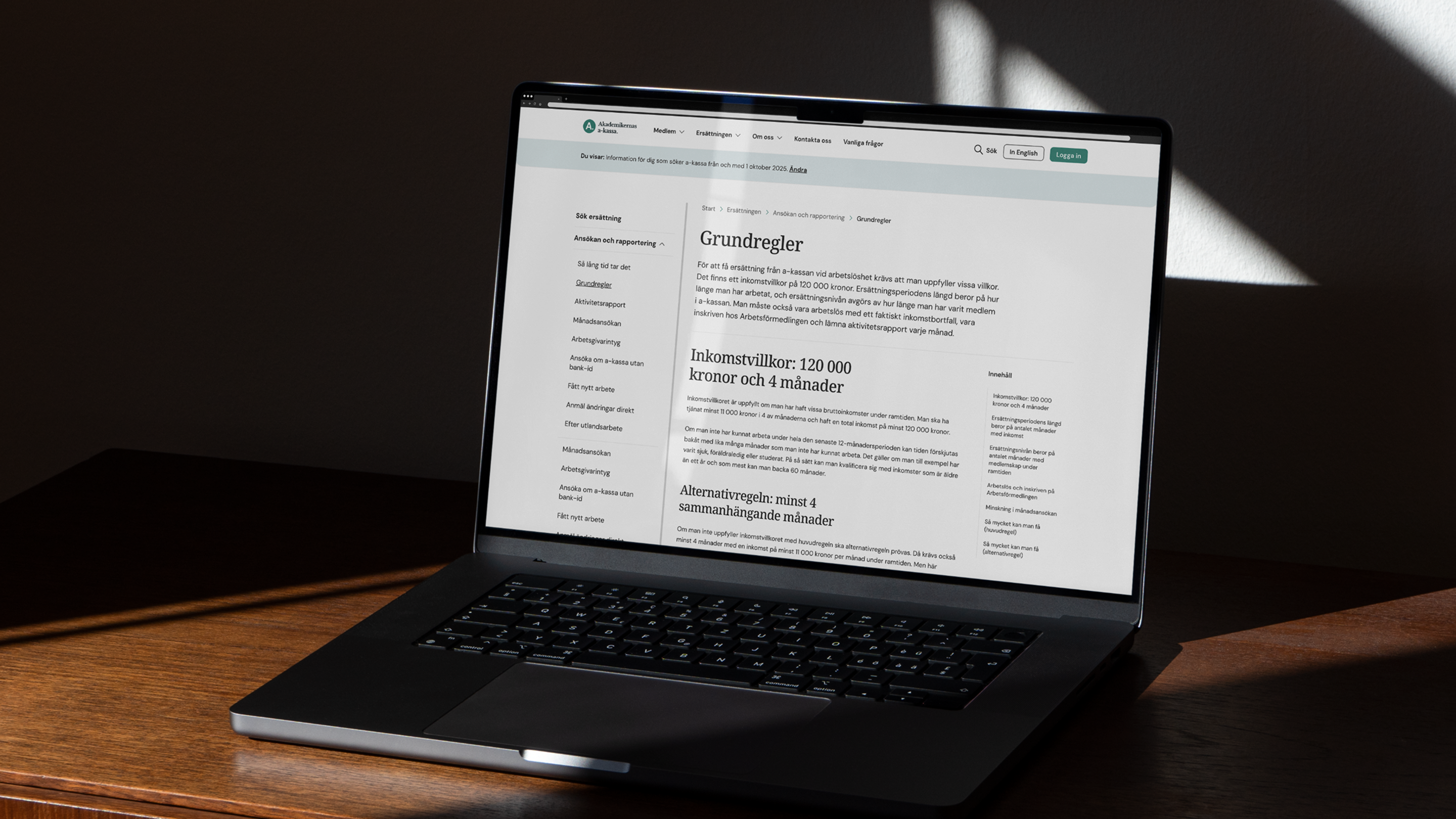 A laptop resting on a wooden desk displaying a text-focused "Grundregler" (Basic Rules) guidance page on the Akademikernas a-kassa website.