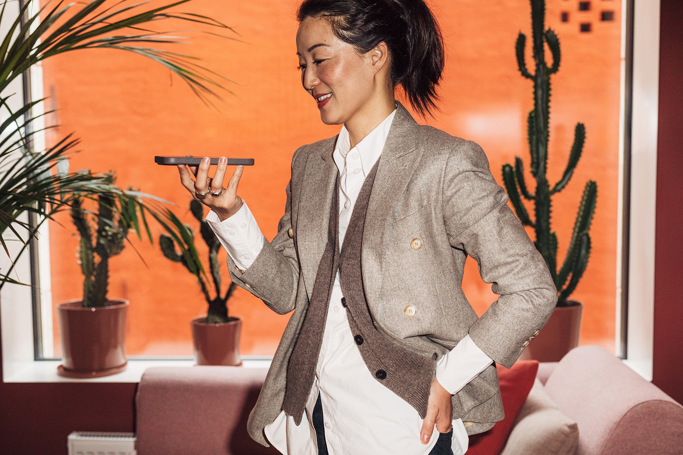 Woman in gray blazer holding a smartphone, smiling while standing indoors near potted cacti on a couch.