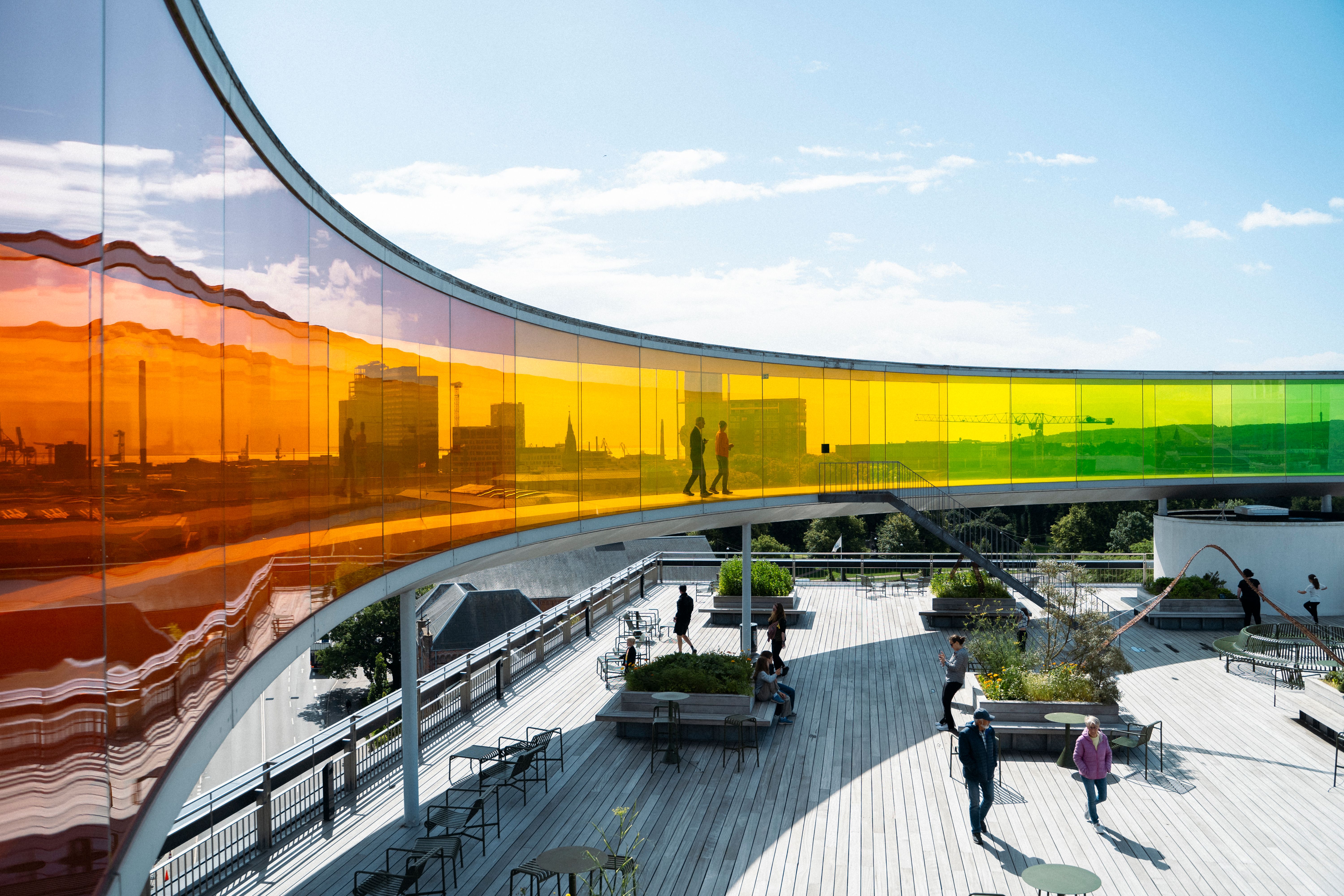 Outdoor terrace beside a curved glass wall with colorful cityscape mural reflections and people walking