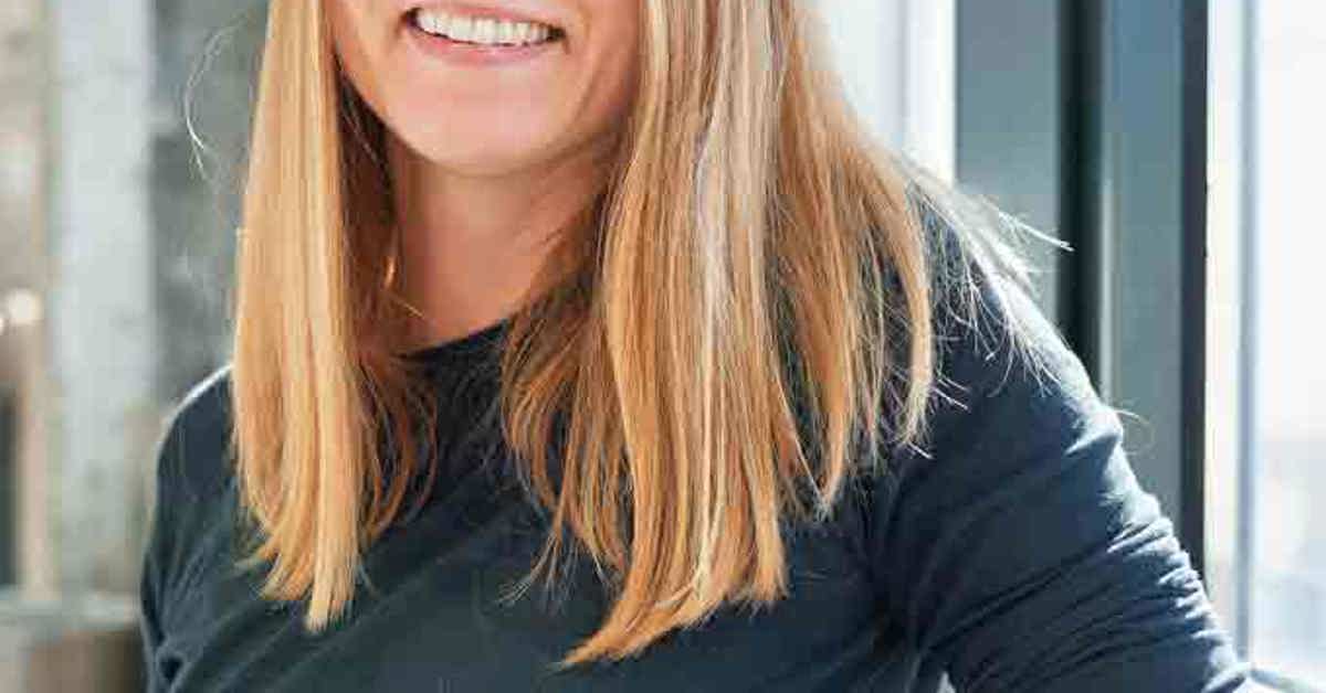 Profile Picture of Erica Quessenberry. A smiling woman with long strawberry blond hair, wearing a grey sweater. She is seated in a brightly lit environment leaning against a window.