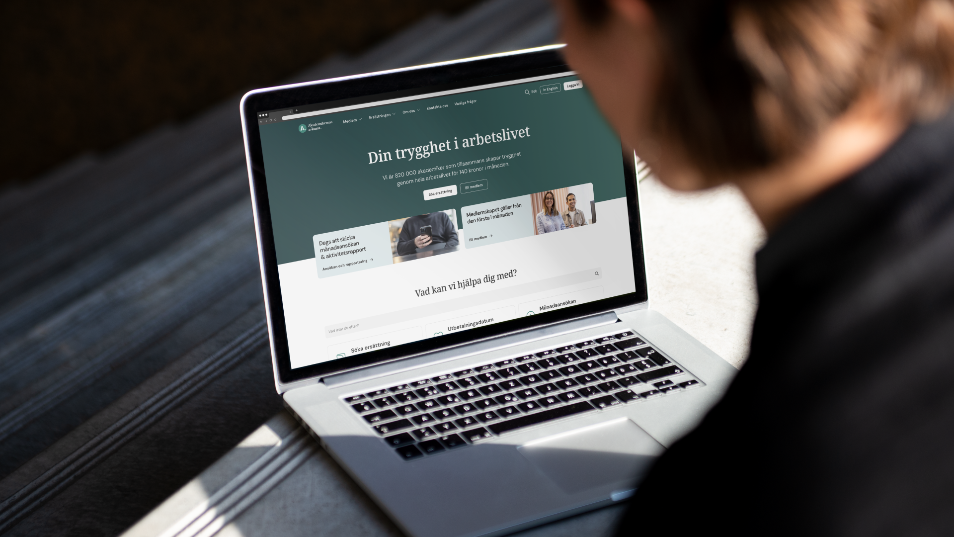 An over-the-shoulder photo of a person using a laptop displaying the Akademikernas a-kassa homepage with the Swedish headline "Din trygghet i arbetslivet".