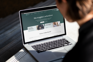 An over-the-shoulder photo of a person using a laptop displaying the Akademikernas a-kassa homepage with the Swedish headline "Din trygghet i arbetslivet".
