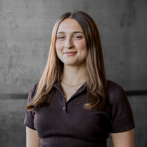 Smiling woman with long light-brown hair wearing a dark polo shirt against a neutral gray background